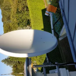 Biała antena satelitarna z pomarańczowym konwerterem LNB zamontowana na dachu, widok z góry na tle zieleni i błękitnego nieba.