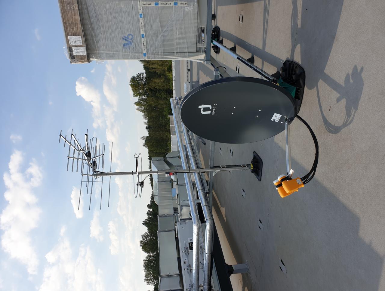 Antena satelitarna Invertlo oraz antena kierunkowa Yagi-Uda zamontowane na płaskim dachu budynku przemysłowego, widoczne kable i złącza, w tle błękitne niebo z chmurami i zalesiony teren.