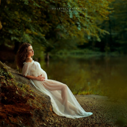 Patrycja Tatarczy Photography - Kobieta w zwiewnej białej sukni, oparta o drzewo nad brzegiem jeziora, w trakcie sesji ciążowej.