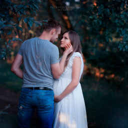 Patrycja Tatarczy Photography - Przyszła mama w białej sukience z falbankami, czule obejmowana przez partnera w plenerze z zielonym tłem.