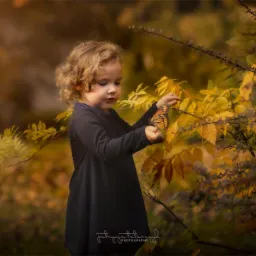 Dziewczynka z kręconymi włosami trzyma motyla monarchę na tle jesiennych liści w ciepłych odcieniach.