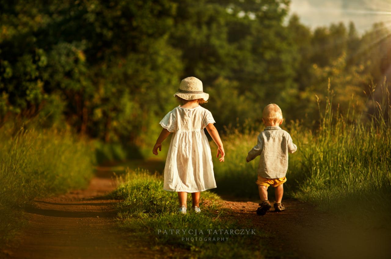 Dziewczynka w białej sukience i słomkowym kapeluszu oraz chłopiec w szarej koszuli i żółtych spodenkach idą polną drogą w stronę lasu, oświetloną promieniami zachodzącego słońca.