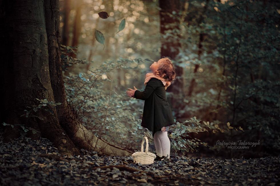 Dziewczynka w ciemnym płaszczu i z koszykiem spogląda w górę na spadające liście w leśnej scenerii, subtelne oświetlenie i stonowana kolorystyka.