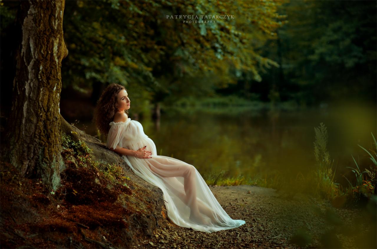 Kobieta w zwiewnej białej sukni, oparta o drzewo nad brzegiem jeziora, w trakcie sesji ciążowej.
