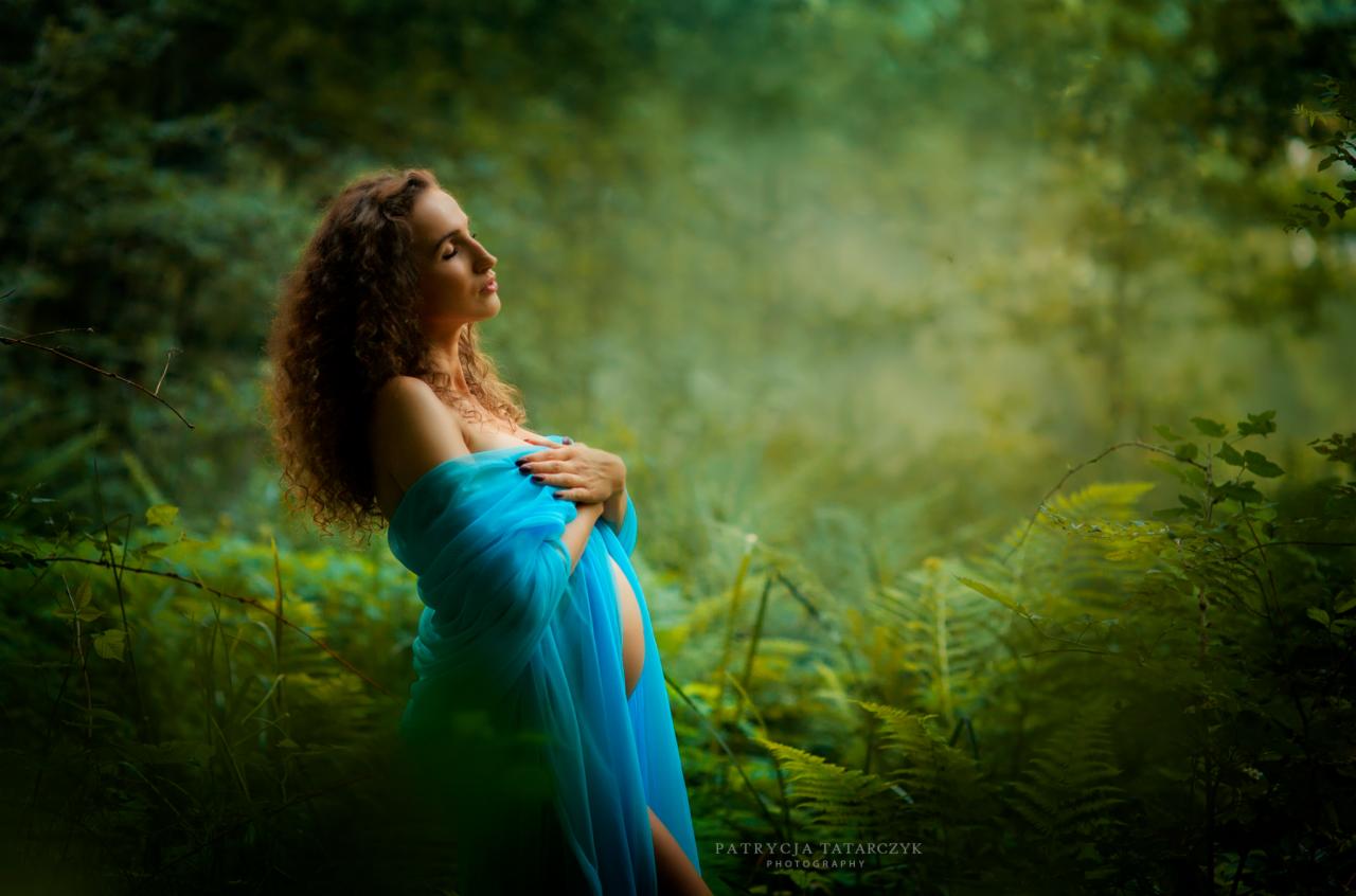Kobieta w ciąży, owinięta błękitnym materiałem, pozuje w zalesionym otoczeniu, z delikatnym światłem padającym na jej twarz.