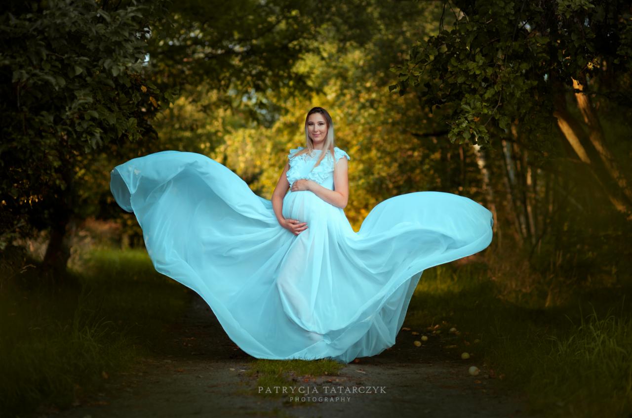 Kobieta w ciąży, w zwiewnej, błękitnej sukni, pozuje na leśnej ścieżce, z delikatnym uśmiechem i dłońmi na brzuchu.
