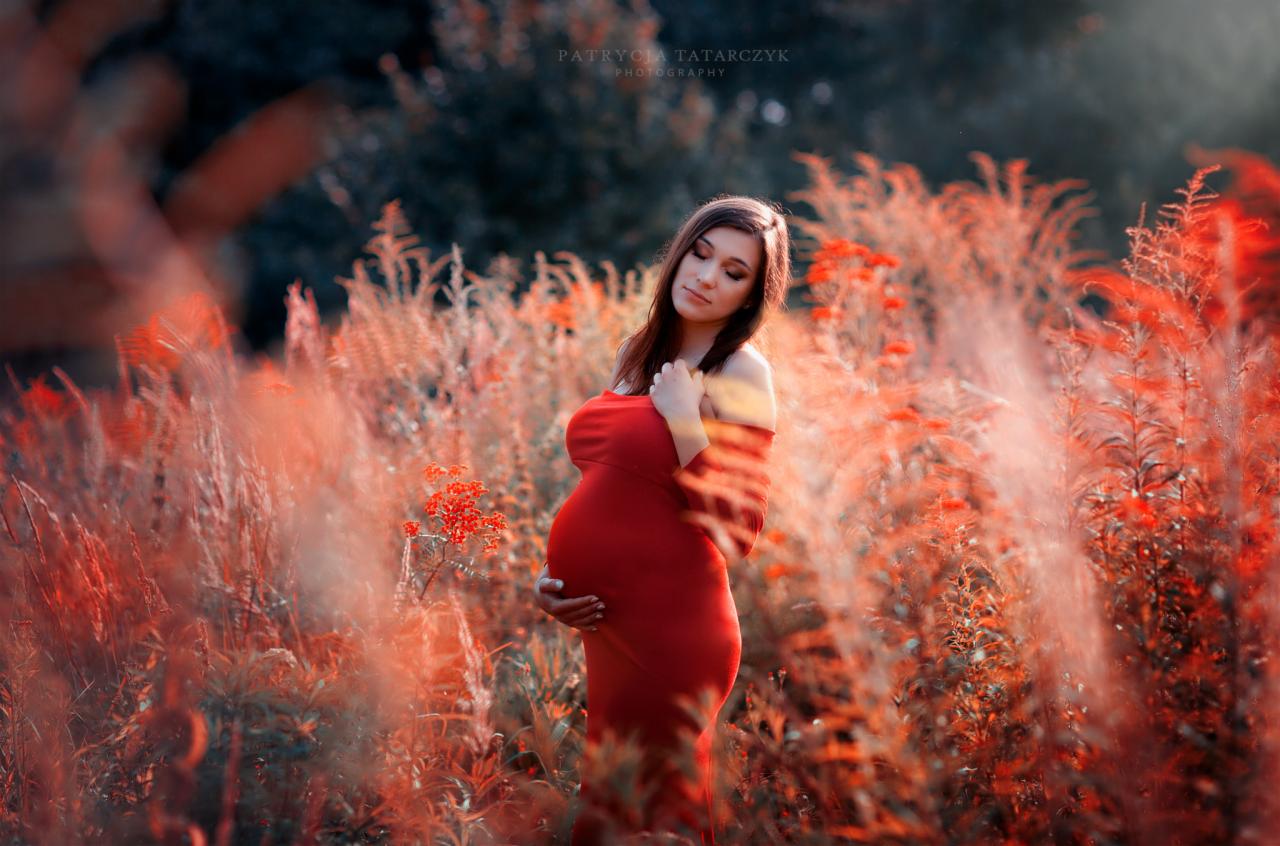 Kobieta w czerwonej sukni ciążowej pozuje w plenerze wśród pomarańczowej roślinności, delikatne światło słoneczne.