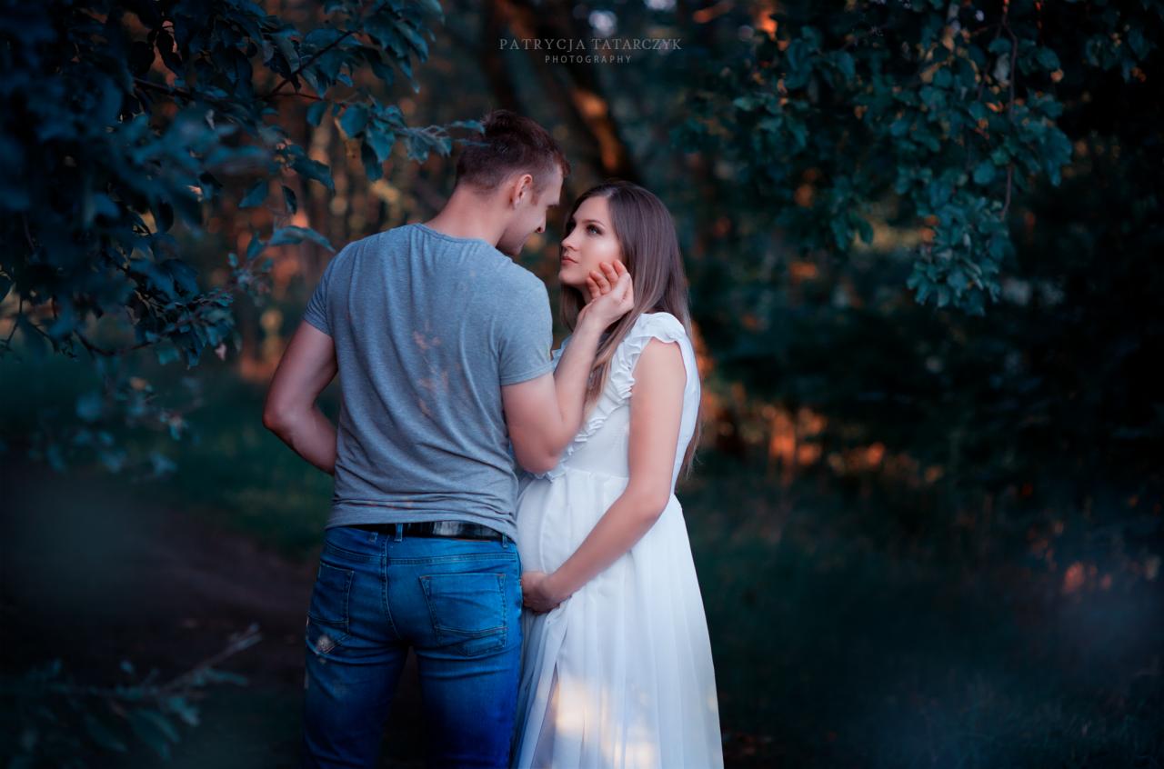 Przyszła mama w białej sukience z falbankami, czule obejmowana przez partnera w plenerze z zielonym tłem.