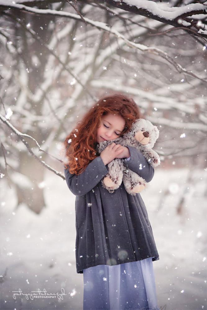 Rudowłosa dziewczynka w szarym płaszczu przytula pluszowego misia na tle zimowego krajobrazu podczas opadów śniegu.