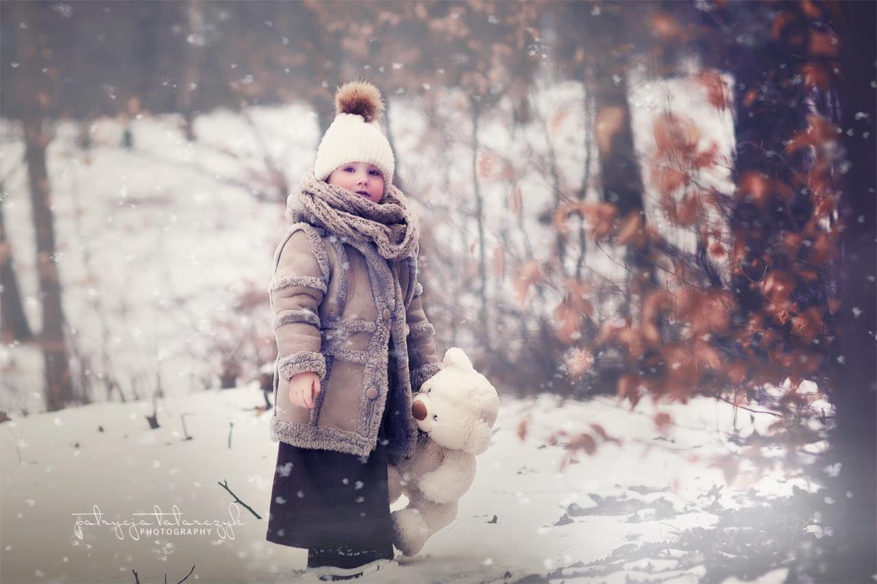 Dziewczynka w zimowym ubraniu z pluszowym misiem na tle zasypanego śniegiem lasu, podczas padającego śniegu.