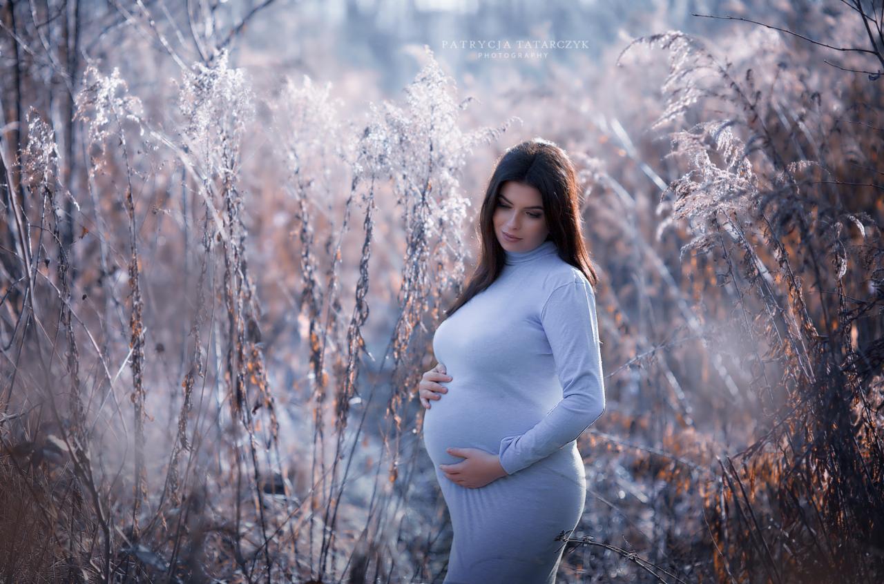 Kobieta w ciąży w obcisłej sukience, pozuje w plenerze wśród wysokich traw w chłodnej kolorystyce.