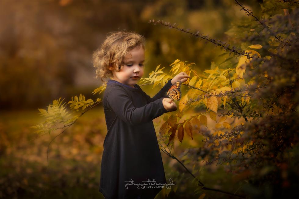 Dziewczynka z kręconymi włosami trzyma motyla monarchę na tle jesiennych liści w ciepłych odcieniach.