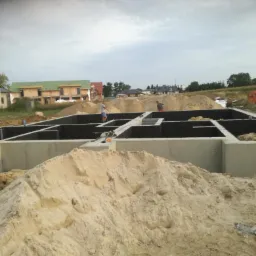 Wylane fundamenty domu jednorodzinnego z izolacją, widoczny kopiec piasku i dwóch pracowników na budowie.