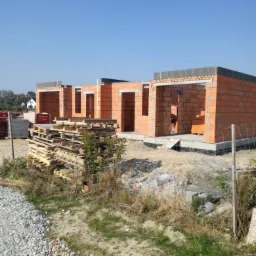 Wznoszenie murów parterowego domu z czerwonej cegły, widoczne palety z materiałami budowlanymi i fragment terenu budowy.