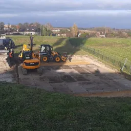 Dwa żółte mini-ładowarki JCB na placu budowy, przygotowujące podłoże z betonowych płyt, w tle widoczne inne maszyny budowlane i hałda gruzu.