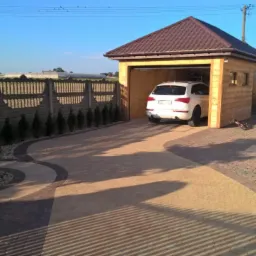 Ułożona z kostki brukowej posesja z drewnianym garażem, betonowym ogrodzeniem i zadbaną zielenią w słoneczny dzień.