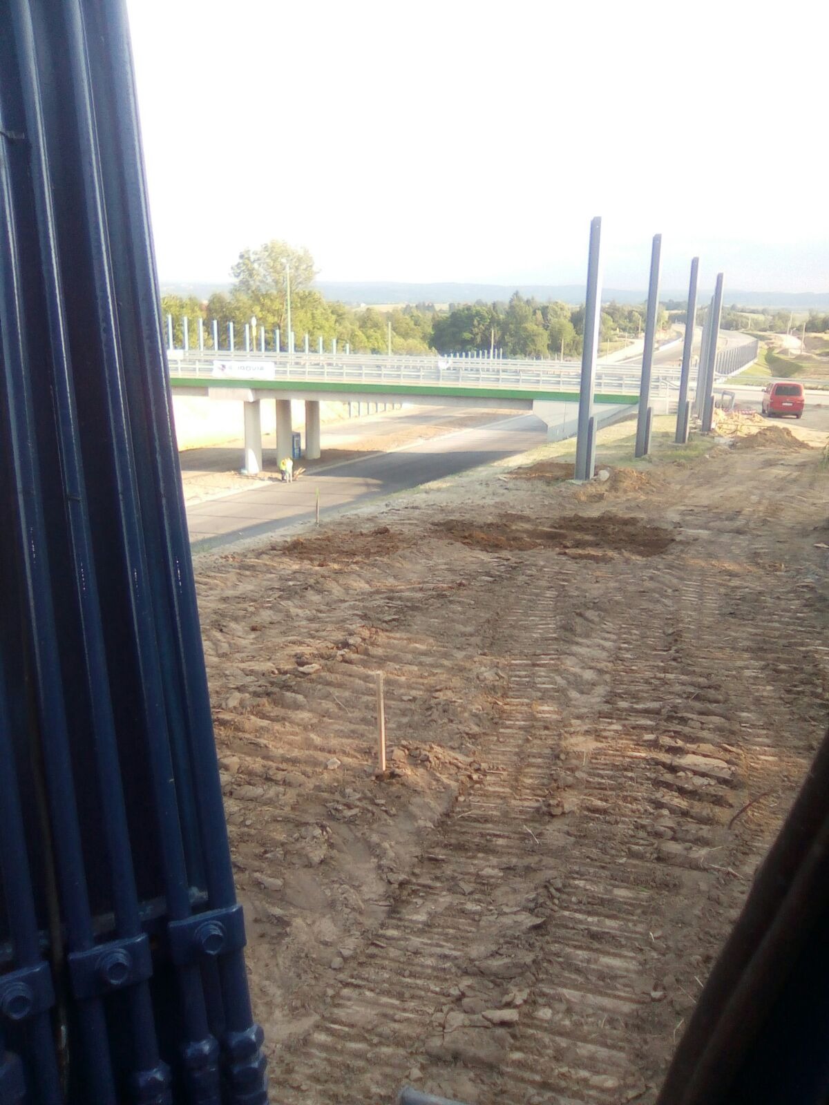 Widok z kabiny maszyny budowlanej na powstający wiadukt z barierami i przygotowany teren pod budowę drogi. W tle zieleń drzew i krzewów, błękitne niebo.