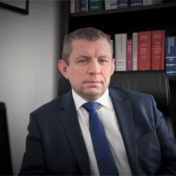 Portret mężczyzny w niebieskim garniturze i krawacie, siedzącego na czarnym krześle biurowym, z regałem pełnym książek w tle.