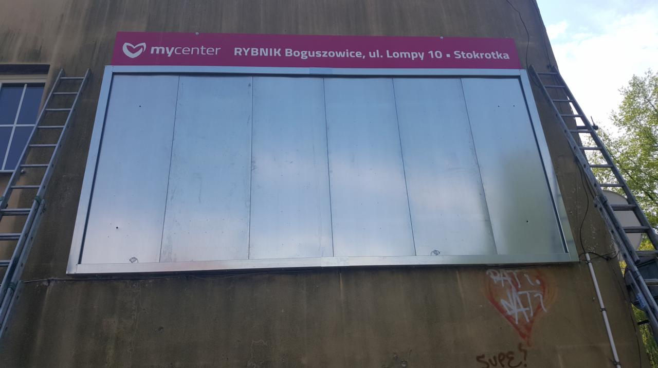 Montaż tablicy reklamowej MyCenter Rybnik Boguszowice na elewacji budynku, widoczne drabiny po bokach i graffiti na ścianie.