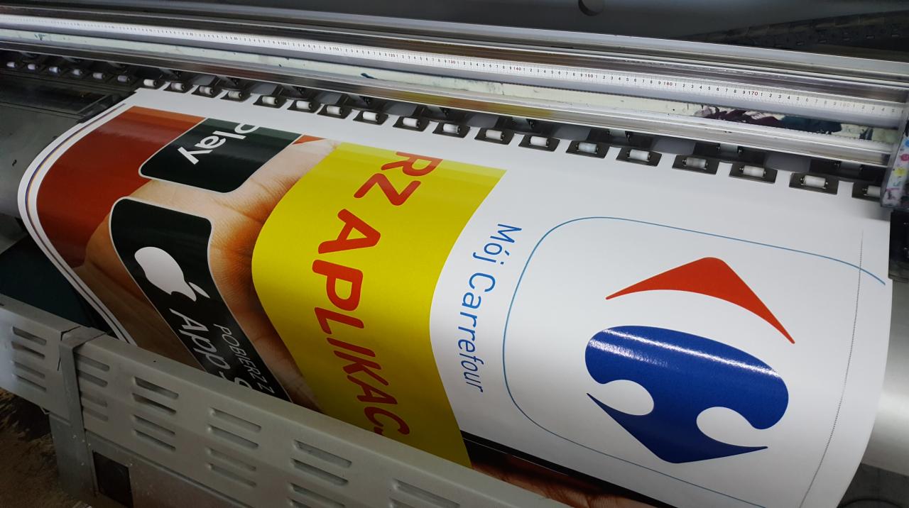 Szeroki wydruk w trakcie realizacji: aplikacja mobilna i logo sieci handlowej na białym tle, widoczne rolki i głowica drukarki.