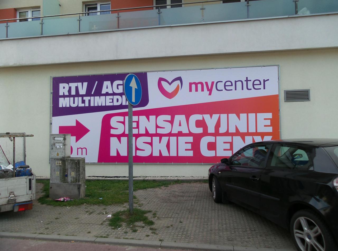 Duży baner reklamowy na ścianie budynku z napisem 'RTV/AG MULTIMEDIA mycenter SENSACYJNIE NISKIE CENY', częściowo zasłonięty przez zaparkowany czarny samochód i znak drogowy.