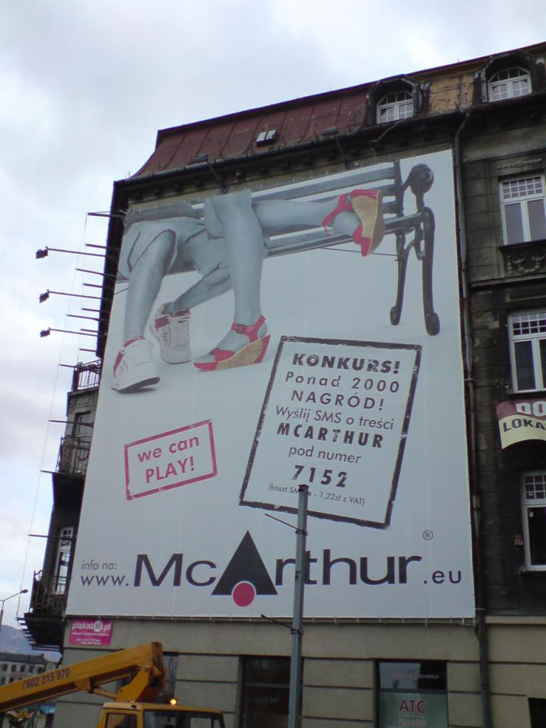 Billboard z grafiką przedstawiającą nogi siedzące na ławce i informacją o konkursie, umieszczony na ścianie budynku w miejskiej scenerii.