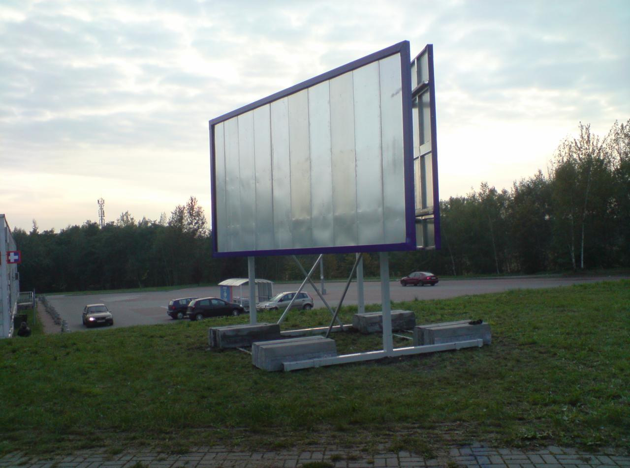 Pusty billboard z fioletową ramą, ustawiony na betonowych blokach, widok na parking, las i pochmurne niebo w tle.