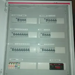 Otwarta skrzynka rozdzielcza z bezpiecznikami i wyłącznikami, opisana etykietami 'RP.3F.INDUKCJA', 'RP.3F.KUCHNIA', 'RP.DWOR', 'RP.LAZIENKI', 'RP.KOTLOWNIA', 'RP.POKOJE', 'OCHRONNIK', 'SYG.FAZ'...