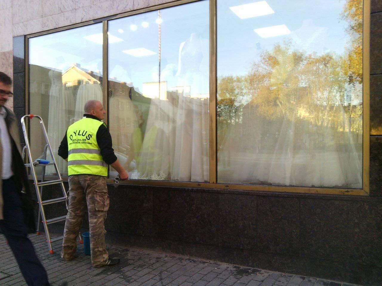 Pracownik w kamizelce odblaskowej z logo firmy czyści dużą witrynę sklepową z manekinami w sukniach ślubnych, widoczna drabina aluminiowa i wiadro z płynem.