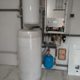 Biały zbiornik buforowy owinięty folią, obok wiszący na ścianie srebrny panel sterowania i niebieski zbiornik wyrównawczy, instalacja grzewcza z szarymi rurami w pomieszczeniu z marmurową podłogą.