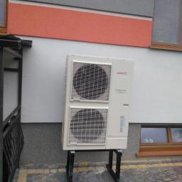 EKOINSTAL POMPY CIEPŁA Jarosław Sobiepanek - Biała jednostka zewnętrzna klimatyzacji marki Atlantic zamontowana na czarnym, metalowym stelażu przy ścianie budynku z elementami w kolorach szarym, pomarańczowym i białym. Widoczny fragment okna...