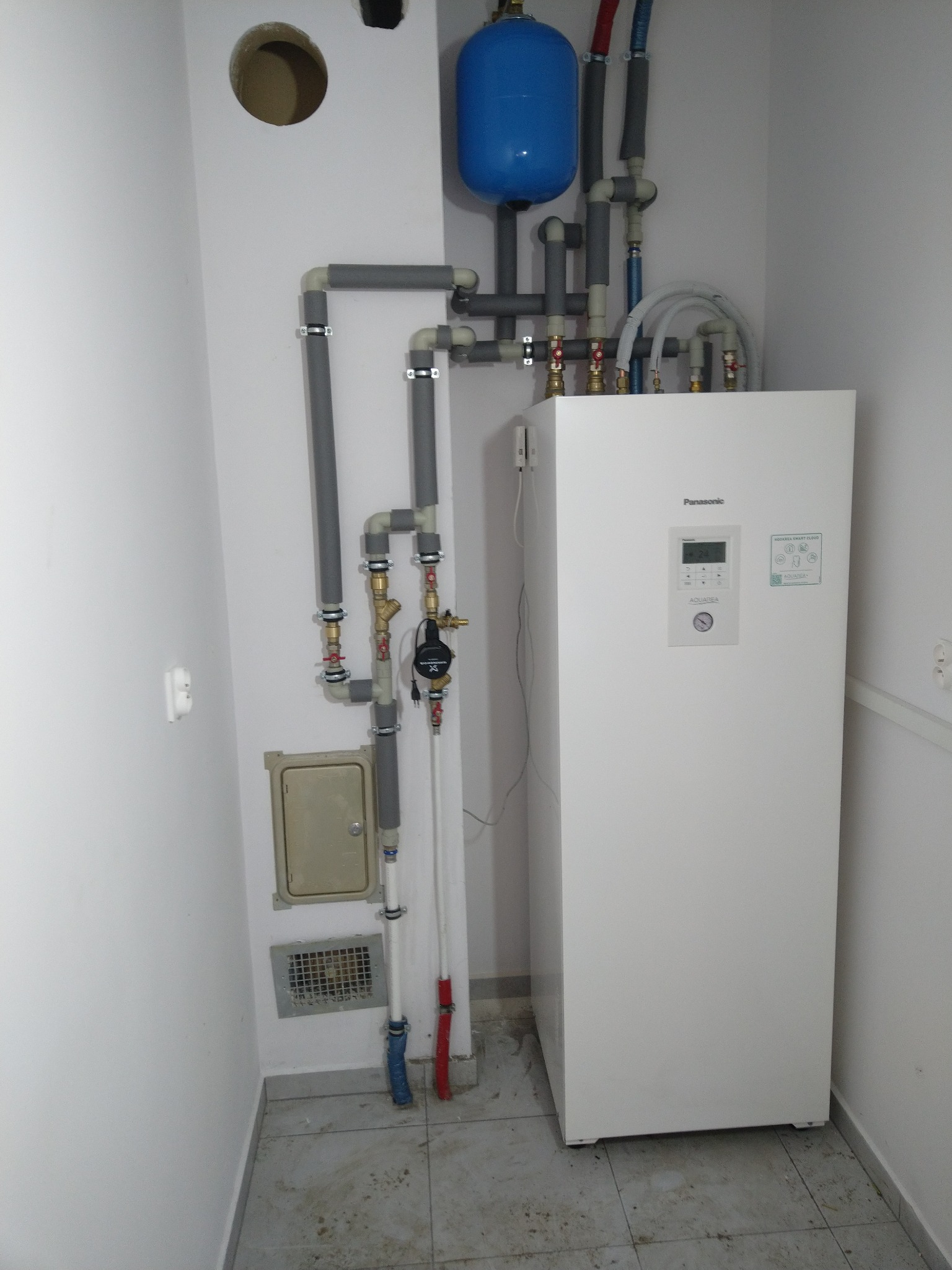 Biała pompa ciepła Panasonic Aquarea Smart Cloud z podłączonymi rurami, zbiornikiem wyrównawczym i osprzętem w pomieszczeniu technicznym z szarą podłogą z płytek.