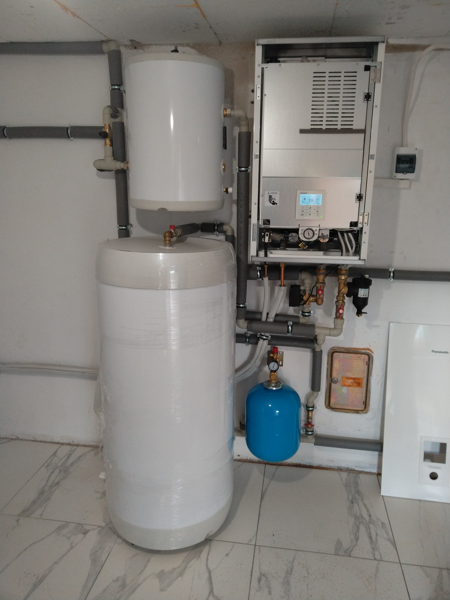 Biały zbiornik buforowy owinięty folią, obok wiszący na ścianie srebrny panel sterowania i niebieski zbiornik wyrównawczy, instalacja grzewcza z szarymi rurami w pomieszczeniu z marmurową podłogą.