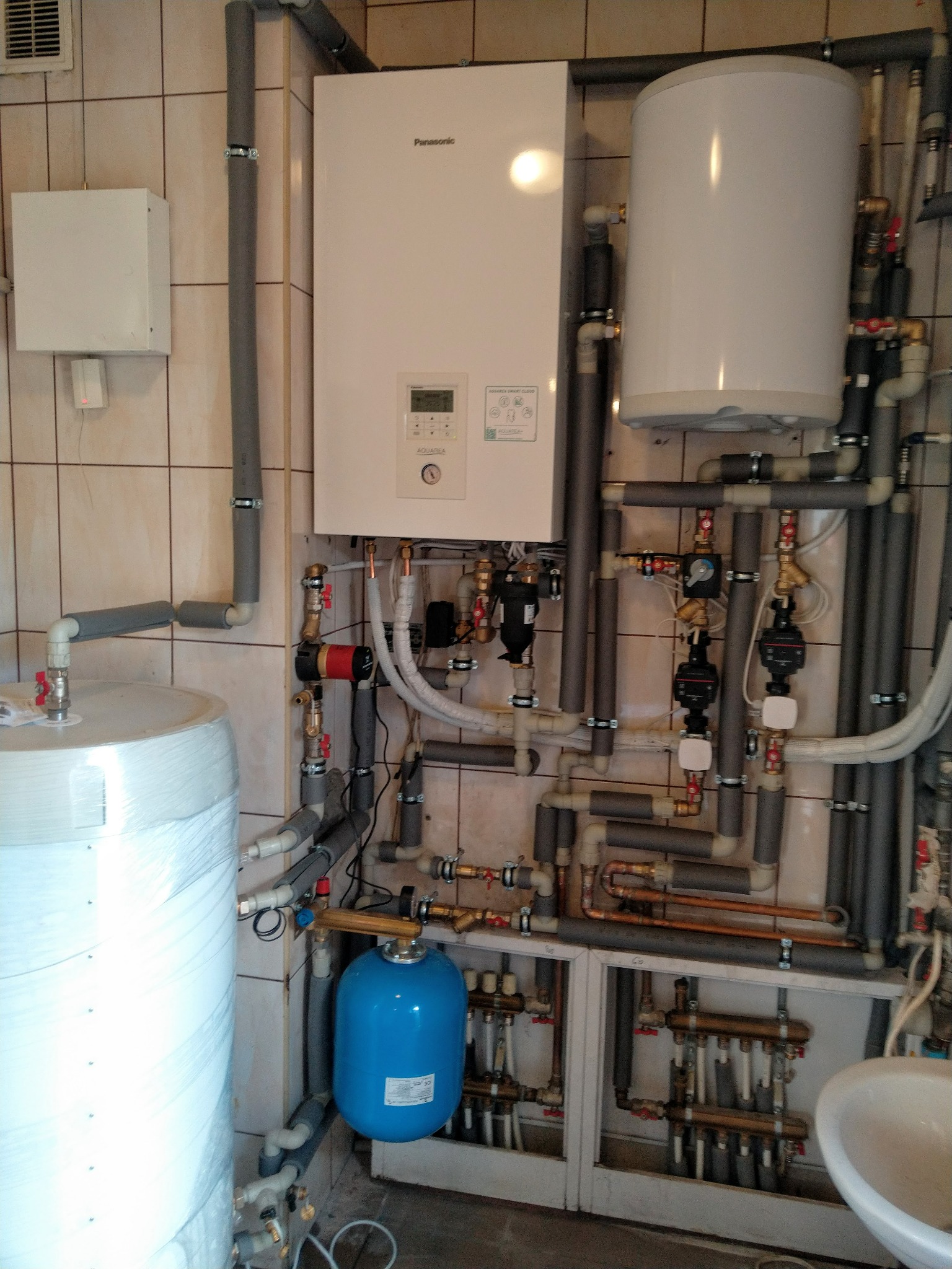 Złożona instalacja grzewcza z białą pompą ciepła Panasonic, zbiornikiem akumulacyjnym owiniętym folią, niebieskim naczyniem przeponowym i rozdzielaczami w szafce, widoczna na tle płytek ceramicznych.