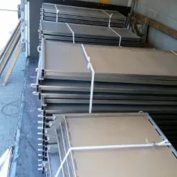 Stalowe elementy konstrukcyjne, związane taśmą, ułożone w stos wewnątrz pojazdu dostawczego. Widoczne otwory montażowe i detale wykończenia.