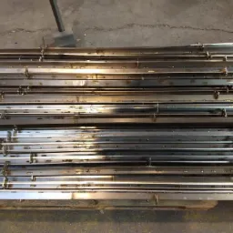 Stos metalowych, spawanych profili o prostokątnym przekroju, ułożonych równolegle na drewnianej palecie, widoczne ślady spawania i otwory montażowe.