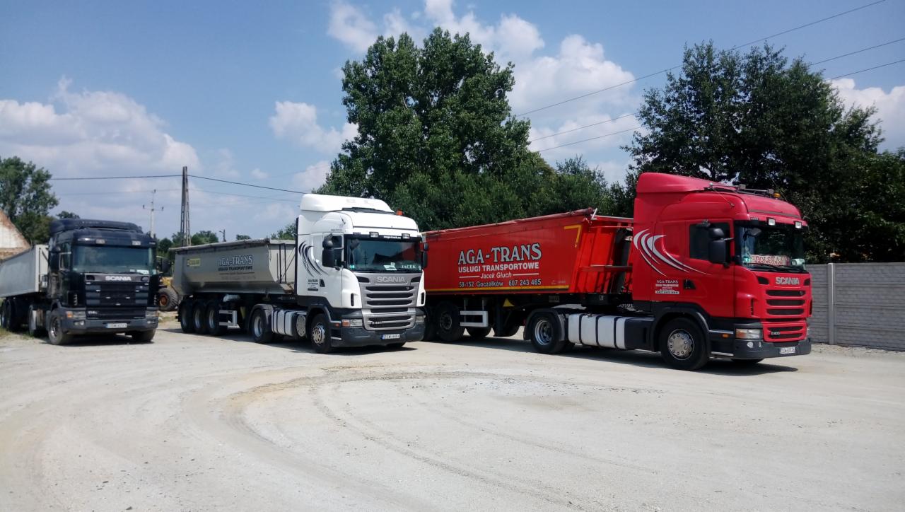 Trzy ciężarówki Scania (czarna, biała i czerwona) z naczepami typu wywrotka, zaparkowane na placu firmy transportowej w słoneczny dzień.