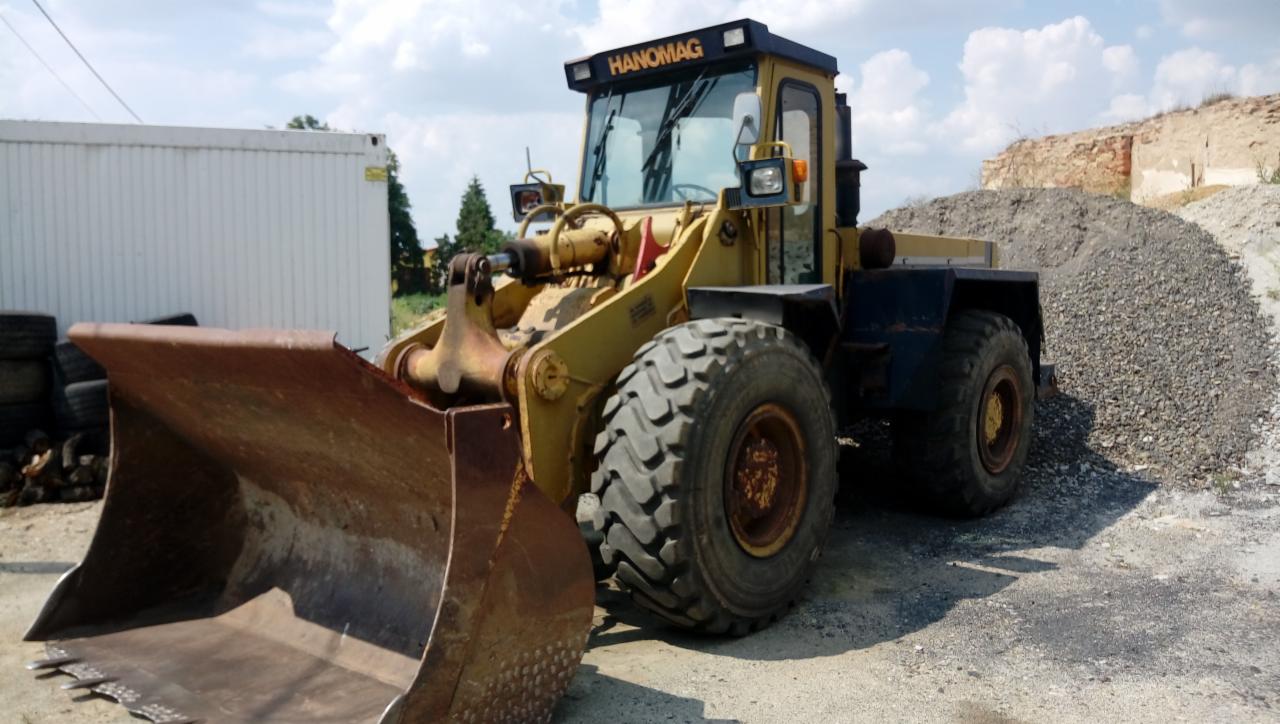Żółta ładowarka kołowa Hanomag z dużym, rdzawym łyżką, stojąca obok hałdy żwiru na placu budowy w słoneczny dzień.
