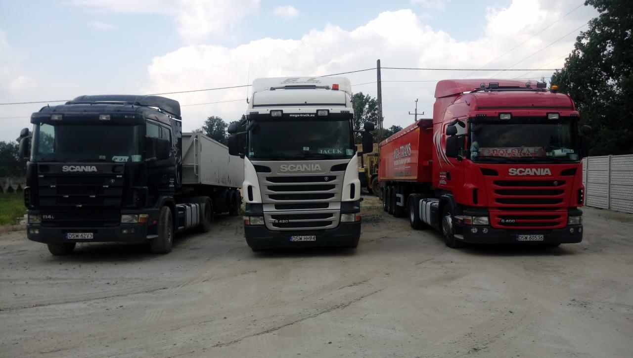 Trzy ciężarówki Scania różnych modeli i kolorów (czarny, biały, czerwony) zaparkowane obok siebie na żwirowym placu w słoneczny dzień.