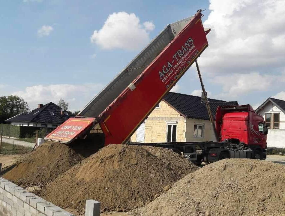 Czerwona ciężarówka wywrotka marki AGA-TRANS w trakcie rozładunku piasku na placu budowy, widoczne domy w tle.