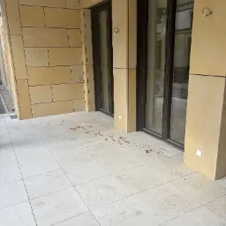 Wykończony taras z jasnych kamiennych płyt kwadratowych, z elementami elewacji z kamienia w odcieniach beżu. Widoczne złącza i elementy montażowe.