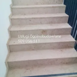 Wykończone schody wewnętrzne z jasnego kamienia z widocznym numerem telefonu firmy budowlanej naniesionym na schody.