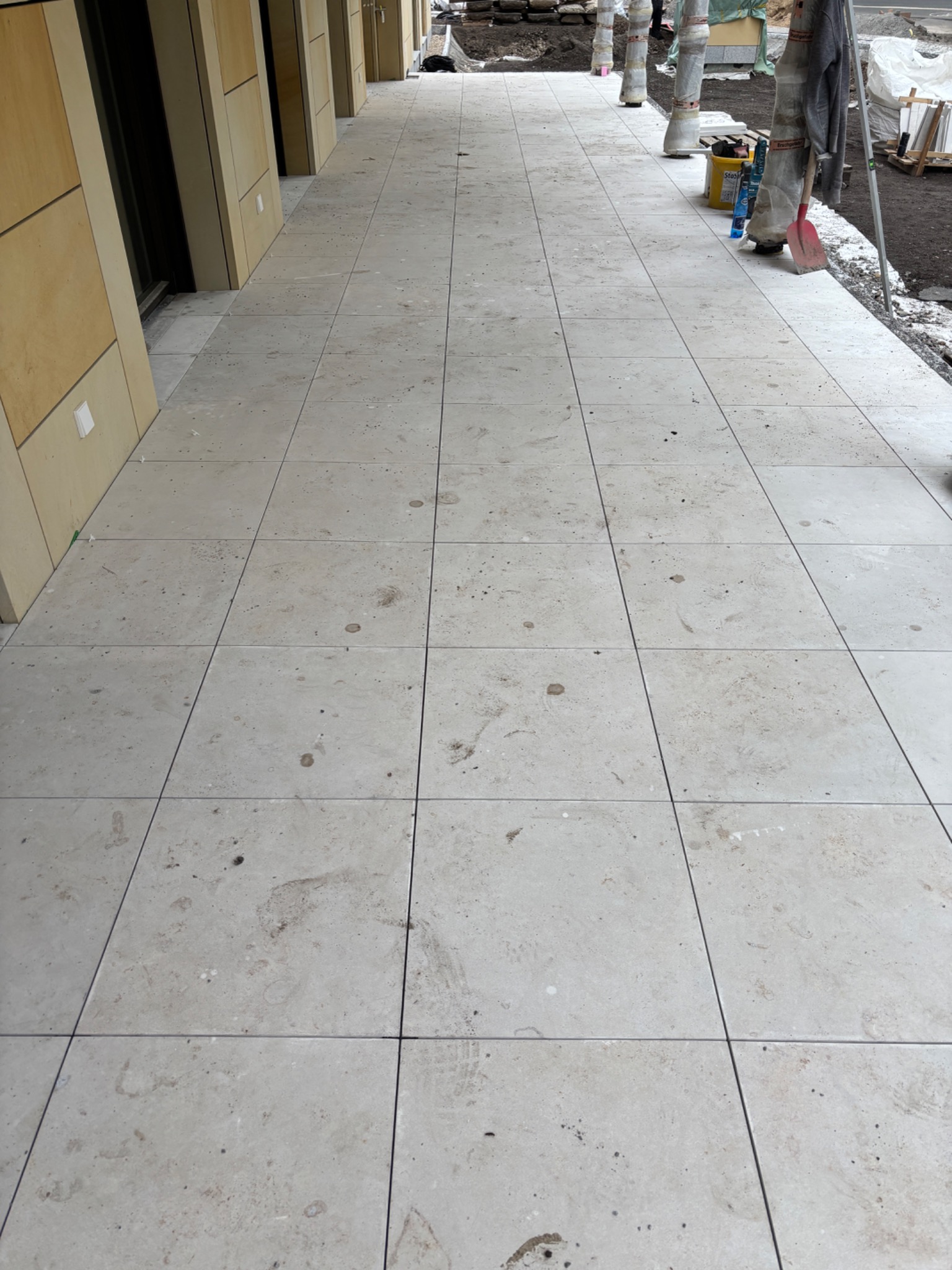 Jasne, kwadratowe płytki tarasowe ułożone na zewnątrz budynku. Widoczne narzędzia i materiały budowlane, sugerujące trwający montaż.