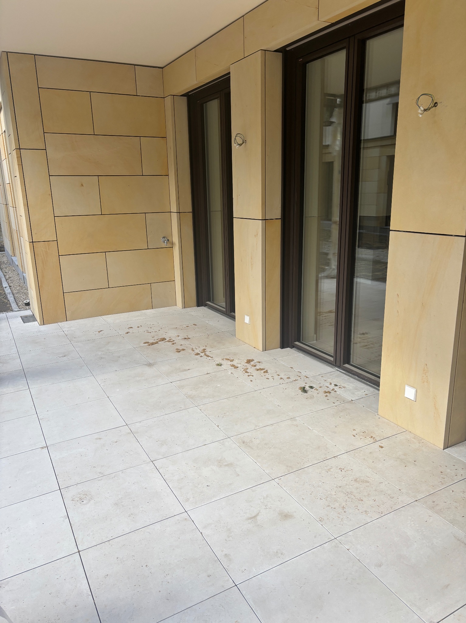 Wykończony taras z jasnych kamiennych płyt kwadratowych, z elementami elewacji z kamienia w odcieniach beżu. Widoczne złącza i elementy montażowe.