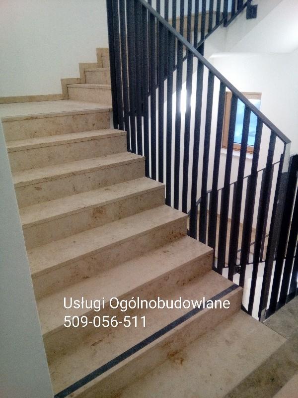 Jasne, kamienne schody z czarną, metalową balustradą wewnątrz budynku, widoczne zabezpieczenia antypoślizgowe na krawędziach stopni.