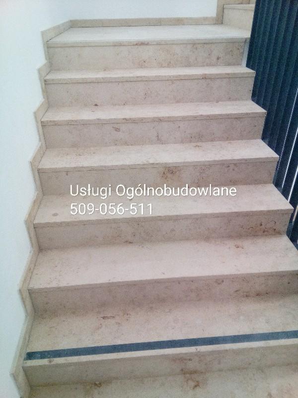 Wykończone schody wewnętrzne z jasnego kamienia z widocznym numerem telefonu firmy budowlanej naniesionym na schody.