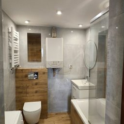 Zbigniew Załęski - Nowoczesna łazienka z szarymi płytkami, drewnianymi akcentami i białym wyposażeniem: umywalka, toaleta, wanna z parawanem i grzejnik. Widoczny piec gazowy.