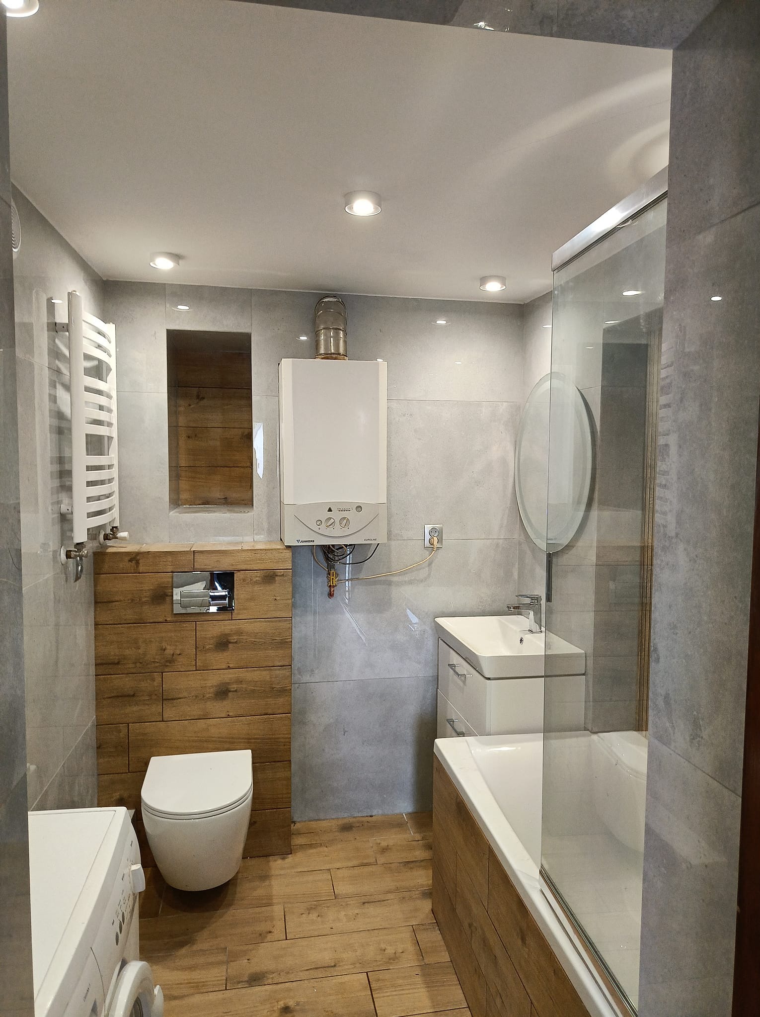 Nowoczesna łazienka z szarymi płytkami, drewnianymi akcentami i białym wyposażeniem: umywalka, toaleta, wanna z parawanem i grzejnik. Widoczny piec gazowy.