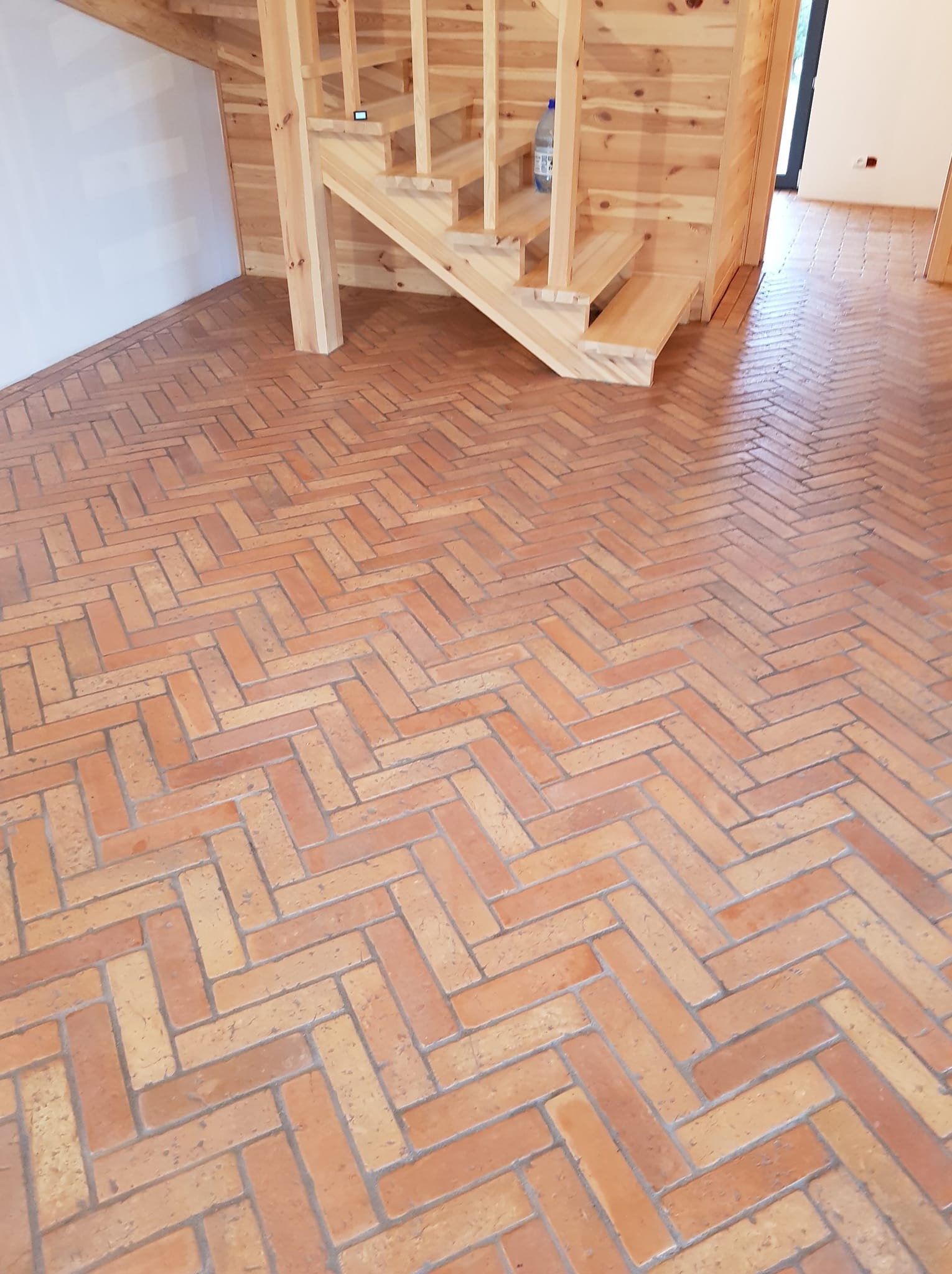 Ukończona podłoga z cegieł ułożonych w jodełkę, widoczna część drewnianych schodów i fragment białej ściany. Styl rustykalny, ciepłe oświetlenie.
