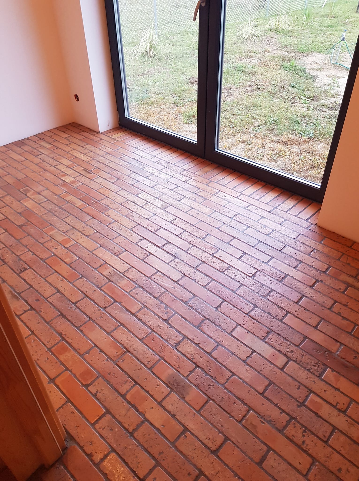 Ceglana podłoga ułożona w jodełkę w jasnym pomieszczeniu z dużym oknem na ogród. Widoczne detale faktury cegieł i gra światła na powierzchni.